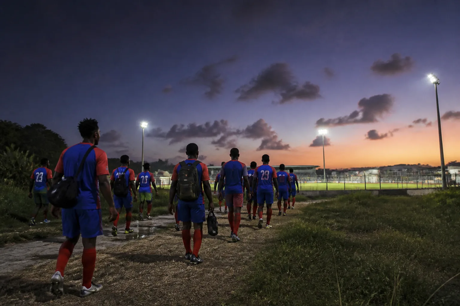 A red-and-blue island football team walking toward a floodlit pitch at twilight