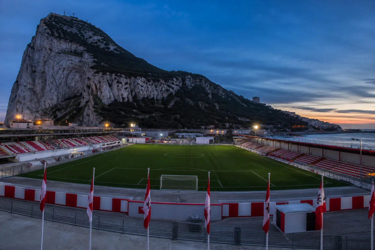 Victoria Stadium in Gibraltar beneath the Rock at blue hour with a clear football pitch and red-and-white flags