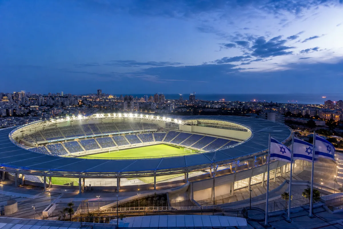 A modern Israeli football stadium at dusk with blue-and-white flags and Mediterranean city lights
