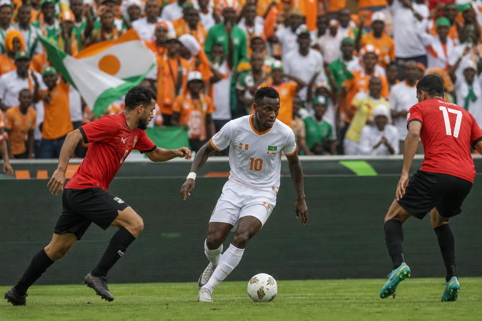 A fictional Niger attacker in white and orange carrying the ball between two North African opponents