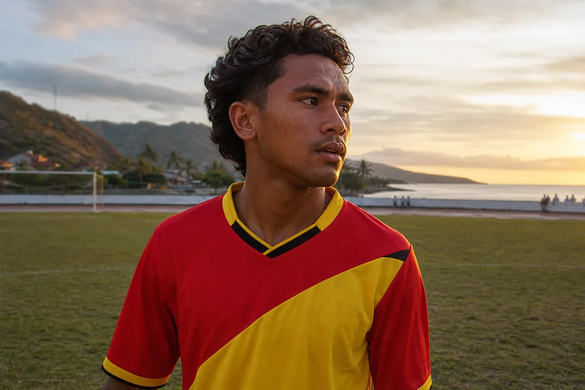 A fictional Timor-Leste national-team attacker inspired by Mouzinho standing on a coastal football pitch near Dili at sunset
